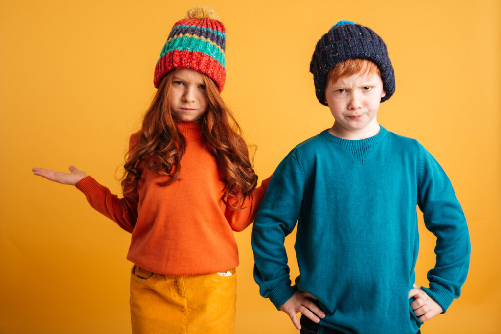 Photo of two confused little redhead children with freckles standing isolated over yellow background wearing warm hats. Looking camera.